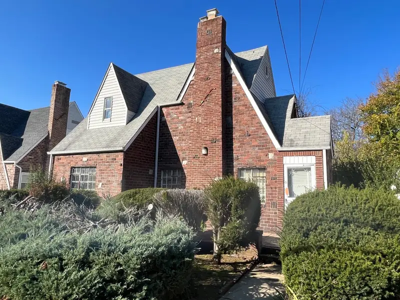 Brick Cape with a tall chimney behind overgrown shrubs, Hempstead, NY